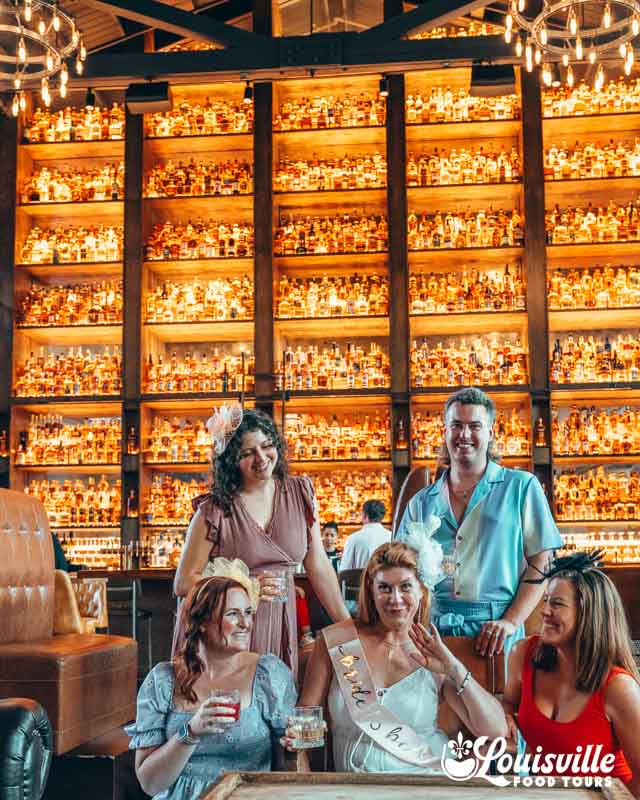 Bride and her bachelorette party in front of a wall of bourbon at the Last Refuge in NuLu
